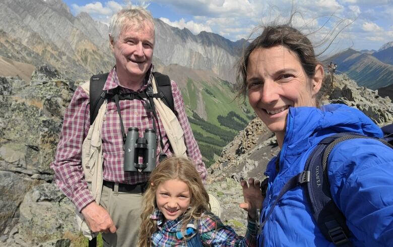 A woman in a blue jacket, a child in a checked shirt and an elderly man in a checked shirt are seen smiling at the top of a mountain with peaks and blue skies in the background.