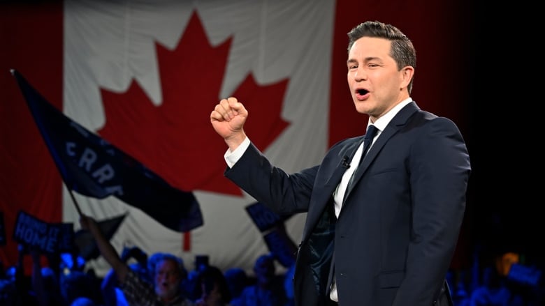 A man in a suit stands on a stage with his fist slightly raised, a Canadian flag hangs in the background.