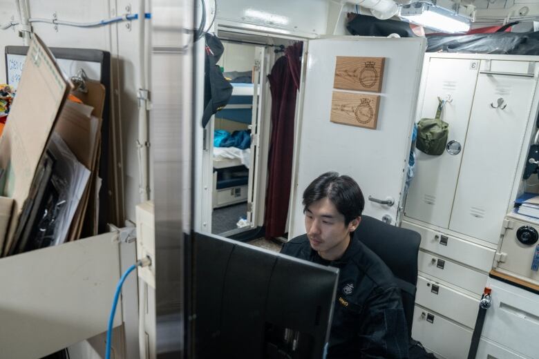 A man works on a computer screen inside a small, crowded room. 