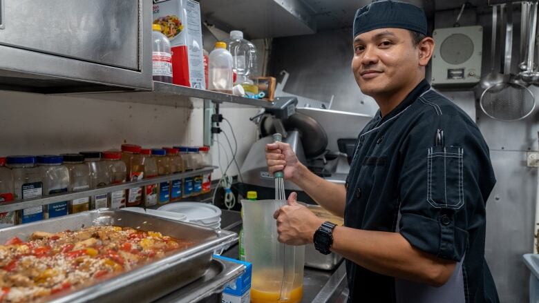 A man wearing an apron and cook's cap smiles while standing in a kitchen area and holding up a jug of sauce that he is stirring. 