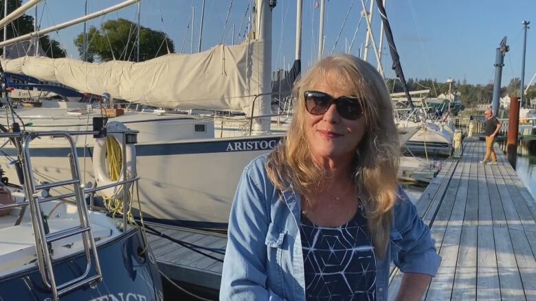 A woman wearing sunglasses and a blue shirt stands on a dock with boats behind her.