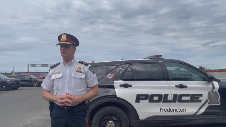 A man with shaved hair in police uniform stands outside, in front of a black and white SUV with Fredericton police force markings.