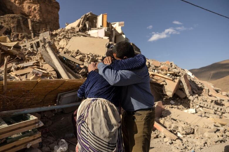 Two people embrace, their heads buried into each other, with rubble visible near them under a blue sky.