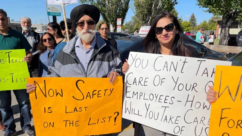 Two people holding signs. A man in a turban holds an orange sign that says 'now safety is last on the list'. A woman standing beside him holds a white sign that says ' you can't take care of your employees how will you take care of us.' 