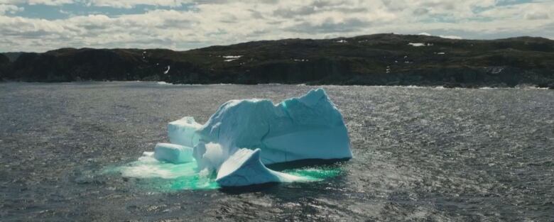 An iceberg floats in the water in the foreground, with a rocky coastline in the background.