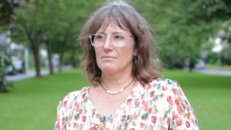 A person with shoulder-length hair, a necklace and glasses looks to the side. Behind her are trees, grass and bushes.