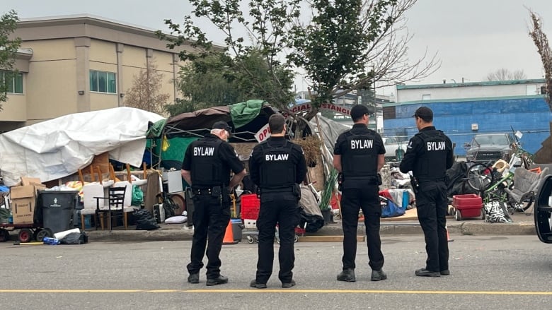 Bylaw officers stand facing a partially demolished encampment