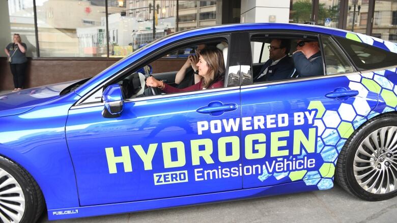 A politician drives a sedan emblazoned with promotion for hydrogen vehicle technology