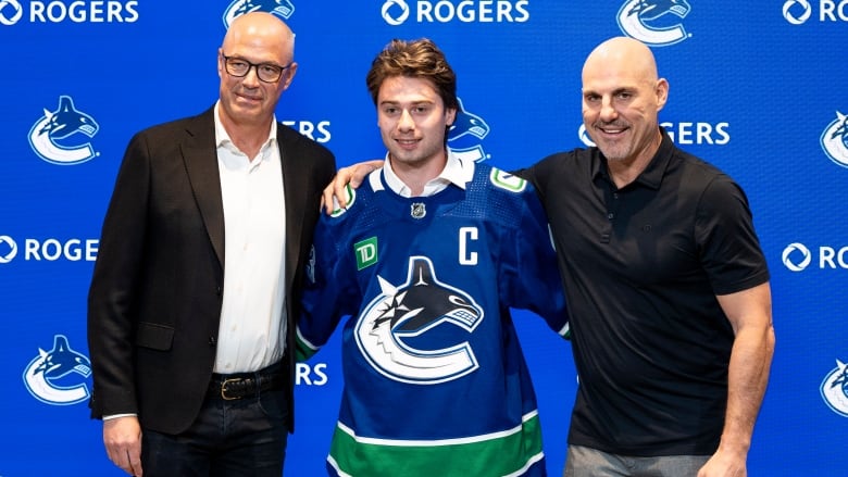 Three men pose for a picture. The one in the centre, younger than the other two, is wearing a blue hockey jersey with a 'C' logo.