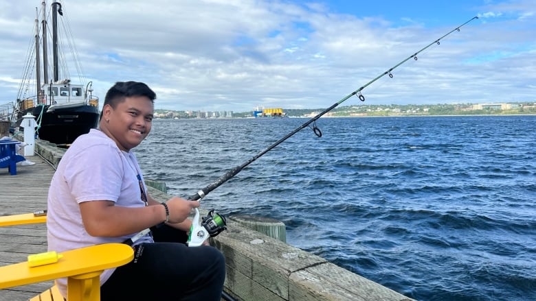 A man sits on a yellow adirondack chair in front of the Halifax harbour. He holds a fishing line. 