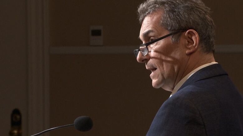 A man wearing reading glasses speaks into a microphone.