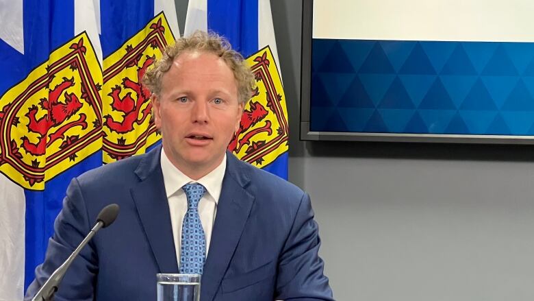 A man in a blue blazer speaks while seated in front of a row of Nova Scotia provincial flags.