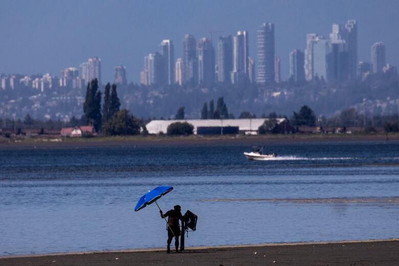 A man holds a blue umbrella above his head on a beach with a city-scape in the background.