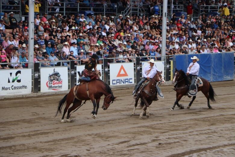 A man rides a horse that is bucking. Two men riding horses look on nearby in the stadium. 