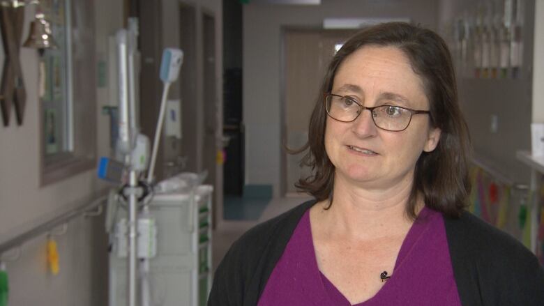 A woman wearing glasses and a purple shirt stands in a hospital hallway.