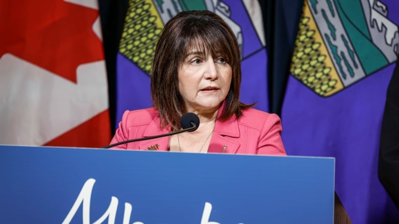 A woman with brown hair speaks at a podium. She is wearing a pink blazer. 