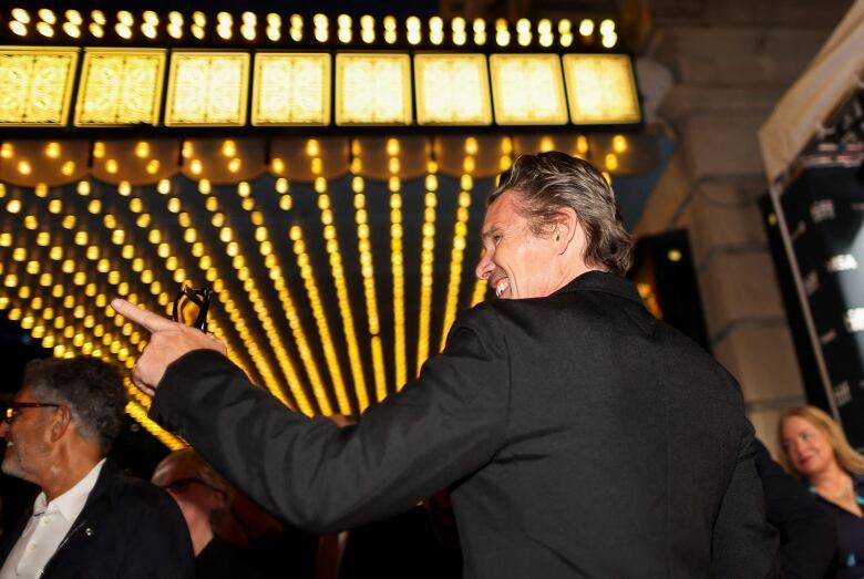 A man in a tuxedo and holding a pair of sunglasses smiles and points while outside a theatre surrounded by people.