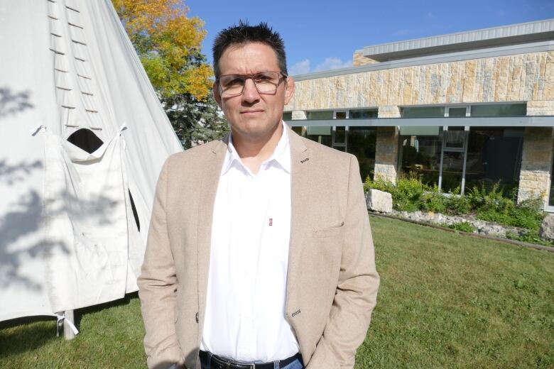 A man with short hair, glasses, a white collared shirt and beige jacket poses for a photo next to a teepee.