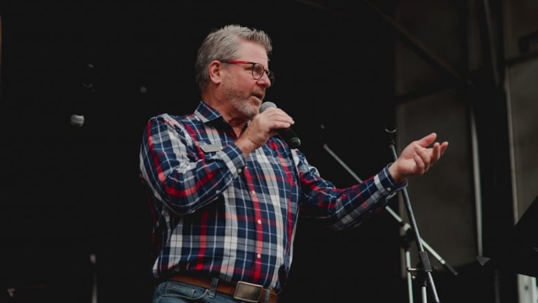 A white man with short grey hair, a beard and glasses stands on a stage in a red, white and blue plaid shirt and jeans, holding a microphone.