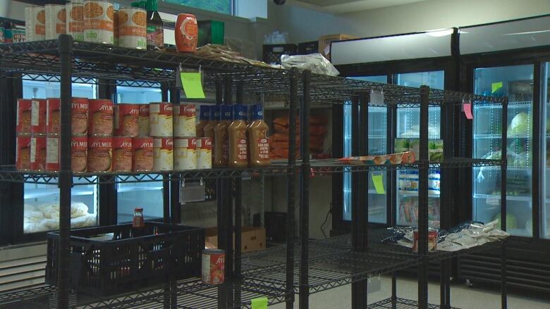 A few metal, black shelves have some canned goods on it. The shelves are mostly empty. In the back, there are some fridges with produce inside. 