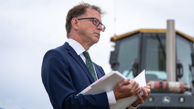 A man in a suit and glasses stands holding an open folder. In the backdrop is a construction vehicle. 