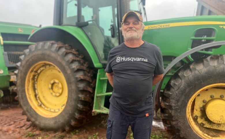 A man with a grey beard stands in front of a large green tractor.