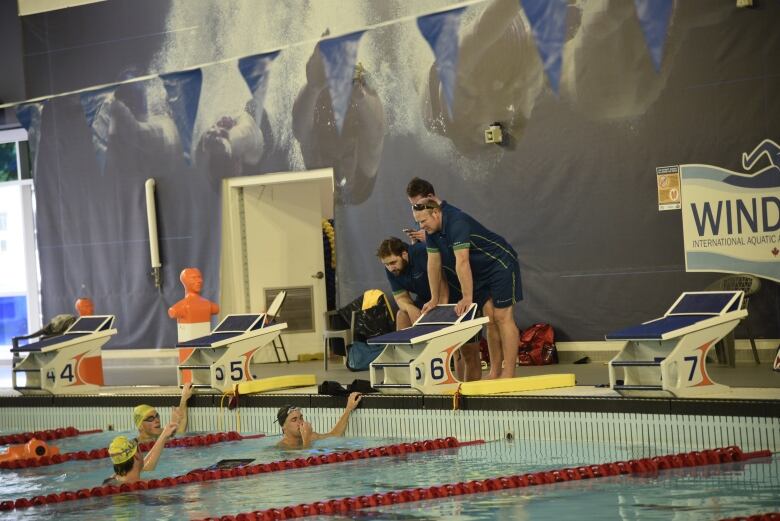 A trio of people in a pool, being spoken to by several people leaning over the edge. 