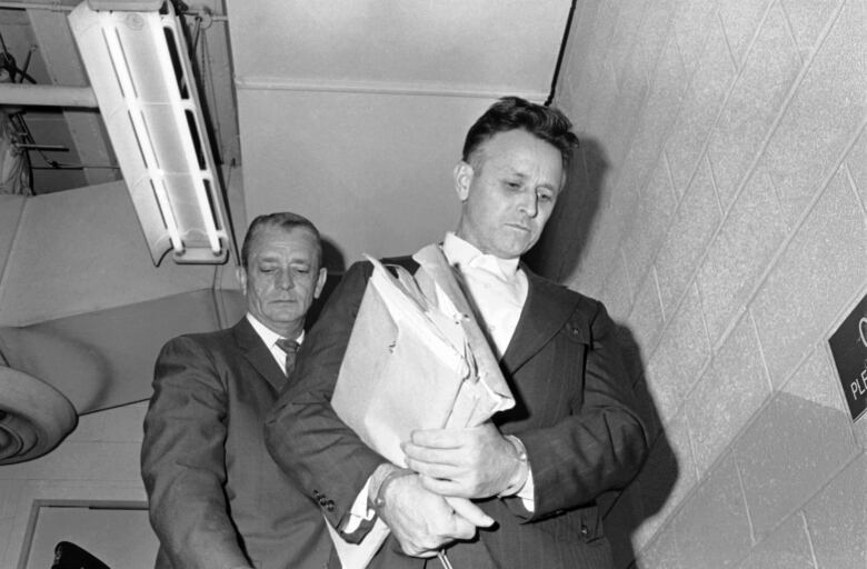 A man is seen holding folders of documents while appearing to walk down a staircase in a black and white photo.