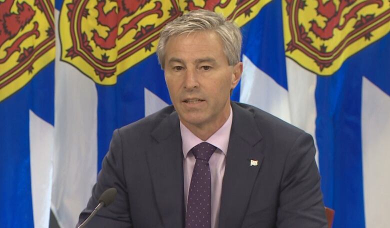 A man in a dark suit, pink shirt and purple tie sits in front of flags.