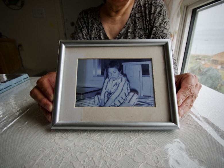 A photo of a young Nancy with short hair, wearing striped pajamas and sitting on a bed. 