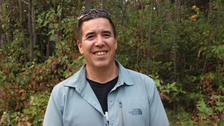 A man standing before forest with a blue shirt and sunglasses on top of his head.