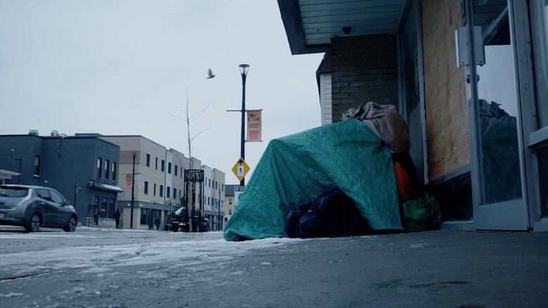 Person under green blanket sits next to a store on the pavement.