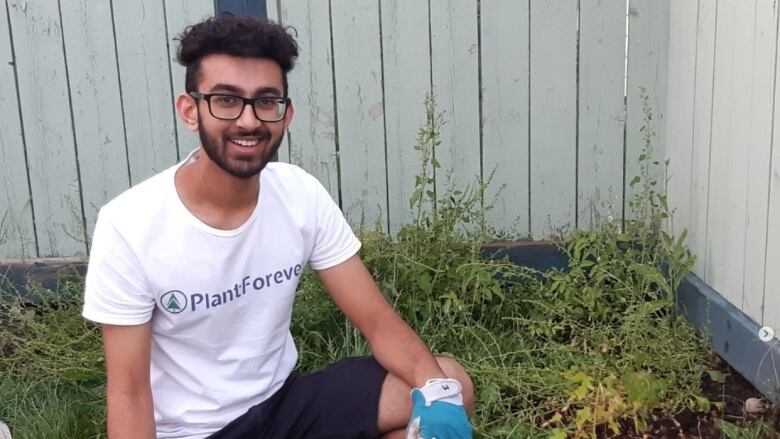 A young man kneels in a house's backyard, next to a freshly planted tree.