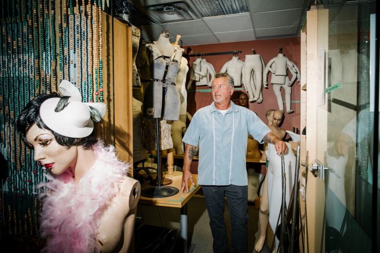 A man with greying hair in a blue shirt and black pants is pictured through a doorway, surrounded by mannequin parts, including a female mannequin in the foreground wearing a pink, feather scarf and a white hat.