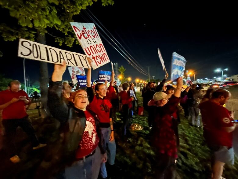 Women and men on an outdoor picket line hold signs, saying 'Solidarity' and 'Detroit is a union town,' during a night time rally.