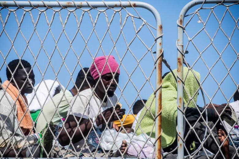 People are shown seated on the deck of a boat, behind a chain-link fence, with their heads wrapped in towels to protect from the sun.