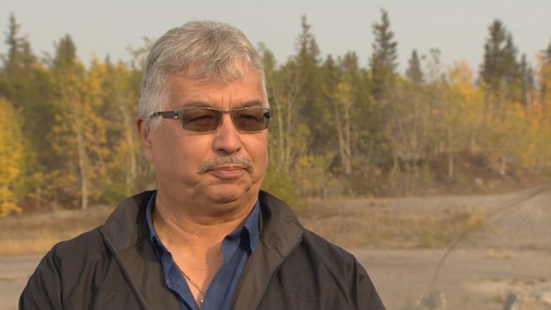 A man wearing glasses and a black jacket stands in front of trees.