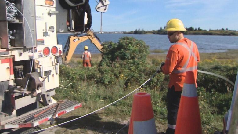 A man wearing a yellow hard hat and an orange shirt holds a wire by a power pole.