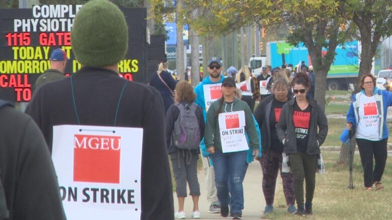 A group of people are seen picketing. One of them has a sign on his back that says 