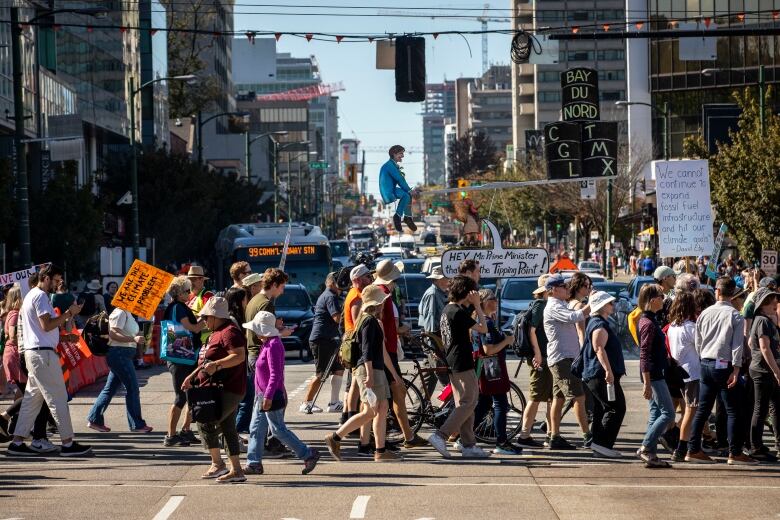 At a busy intersection with cars and a bus in the background, people are crossing the road holding climate change protest signs. 