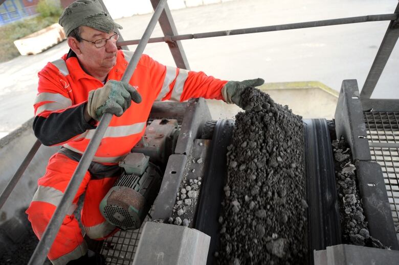 A man wearing a bright orange jumpsuit with reflective bands lets cement clinker from a conveyer belt flow over his gloved hand. 