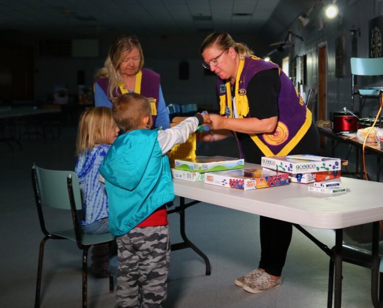 Two children and two women are standing at a table covered in board games in the semi-darkness.