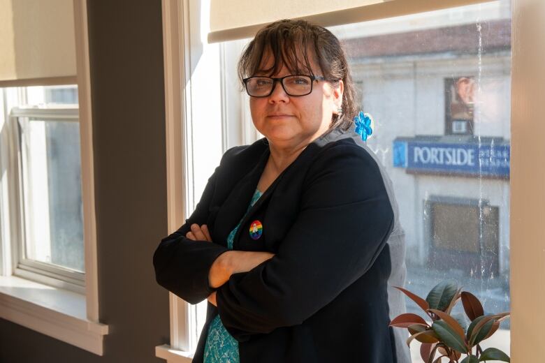 A woman stands against a window with her arms crossed.