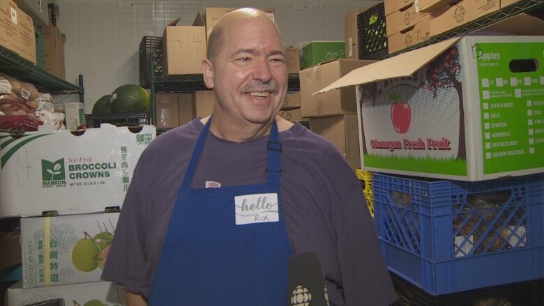 A man in a blue apron smiles inside a crowded walk-in cooler with boxes of fruit and food around him.