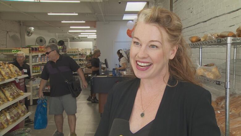 A woman smiles at the camera in a grocery store with bread and goods on shelves behind her.