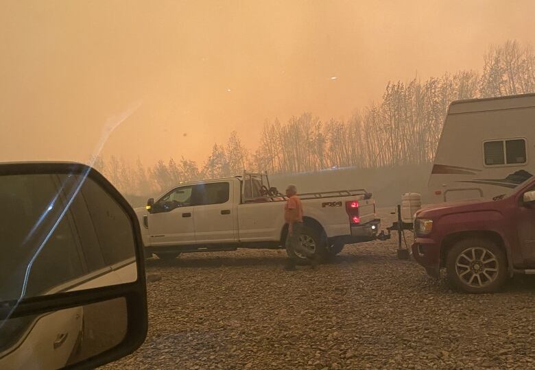 Looking out a vehicle window at a man walking near a pickup truck and camper, parked on rocky ground under an orange, smoky sky.