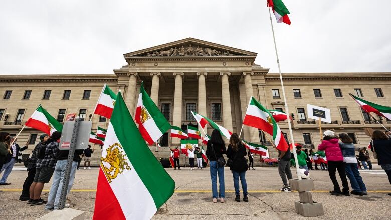 People holding Iranian flags stand in front of a brick building with columns in front of its doorway.