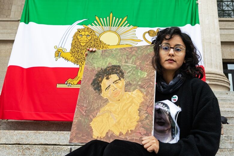 A young women hold a painting of a man while sitting in front of an Iranian flag.