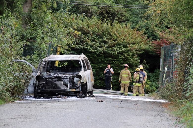 A burned vehicle in a tree-lined area with melted tires and no hood. Firefighters and police stand in the distance behind it.