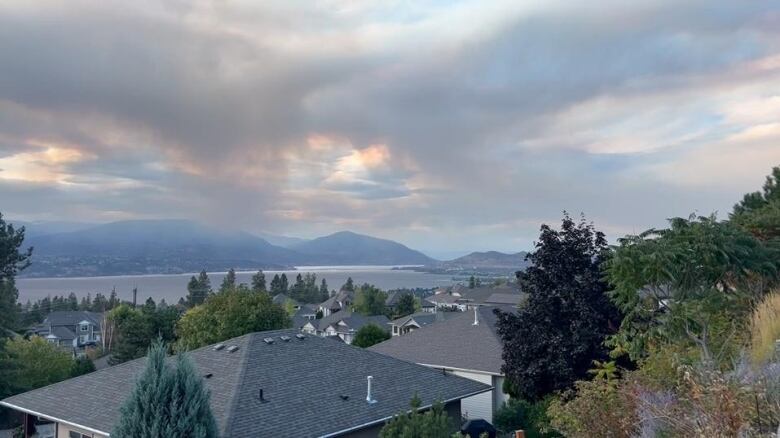 Smoke shrouds a mountain range across a lake from a neighbourhood, with many roofs in the foreground of the picture.